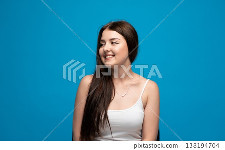 Portrait of a beautiful young woman smiling, feeling joy and confidence, looking sideways on a vibrant blue background Portrait of a beautiful young woman smiling, feeling joy and confidence, looking sideways on a vibrant blue background 138194704
