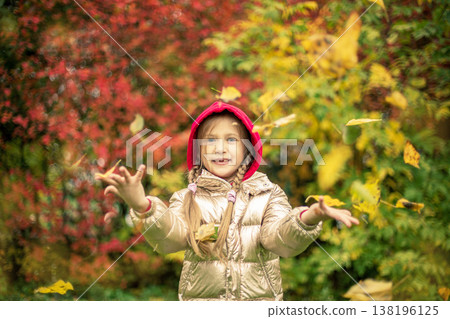 A girl in a gold-colored jacket smiles, throws pink and yellow leaves on an autumn day 138196125