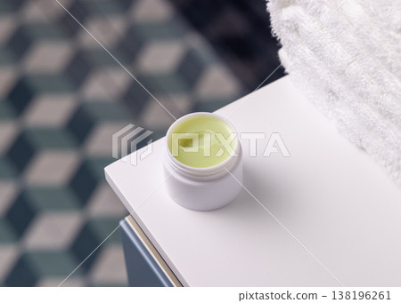 Opened cosmetic green lip balm jar near white bath towel against blue tiled floor, top view Opened cosmetic green lip balm jar near white bath towel against blue tiled floor, top view 138196261