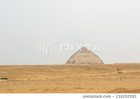 A white pyramid stands in the desert landscape of Dahshur, Egypt. The structure is partially obscured by a hazy sky, surrounded by sandy terrain. 138202091