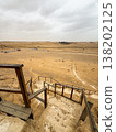 Wooden steps lead down towards a pyramid in a desert landscape. The view from above shows arid terrain and distant structures. 138202125