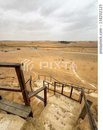 Wooden steps lead down towards a pyramid in a desert landscape. The view from above shows arid terrain and distant structures. 138202125