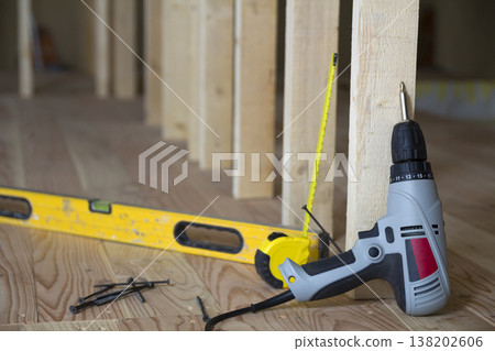 Close-up of professional tools: electrical screwdriver, level and measuring tape on background of wooden frame for future wall in attic room under reconstruction. Renovation and improvement concept. 138202606