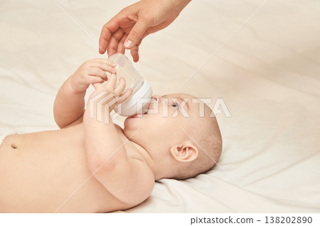 Caucasian Baby Drinking From Bottle With Adult Hand Helping On Soft White Bedding Caucasian Baby Drinking From Bottle With Adult Hand Helping On Soft White Bedding 138202890