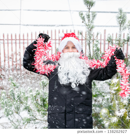 Child dressed as Santa with white beard red tinsel garland standing outdoors in snowy winter garden Child dressed as Santa with white beard red tinsel garland standing outdoors in snowy winter garden 138202927