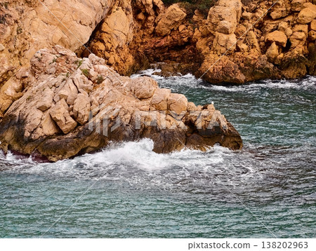 Radiant sunlit rugged rocks meeting frothy waves characterized by dynamic textures and vibrant shades 138202963