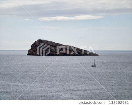Small sailing boat floats near rocky islet, Small sailboat drifts close to rugged island on tranquil sea 138202981