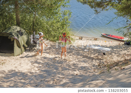 Girls on a sandy beach by the lake shore at a camping site with tents. The water outlet is surrounded by trees. A SUP-boarding is moored on the shoal 138203055