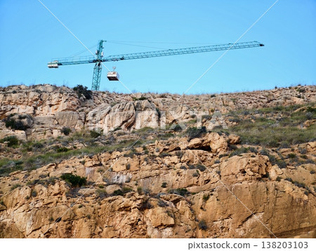 Crane hoists load over ridge, Heavy machinery raises cargo over rocky landscape under open sky 138203103