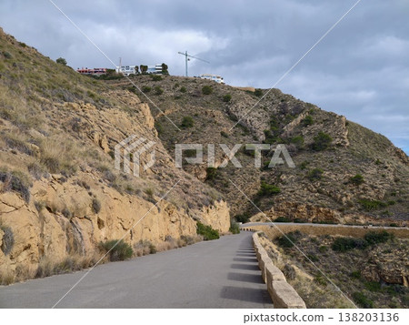 Tranquil asphalt highway following rugged rocky incline under overcast sky with distant scattered buildings Tranquil asphalt highway following rugged rocky incline under overcast sky with distant scattered buildings 138203136