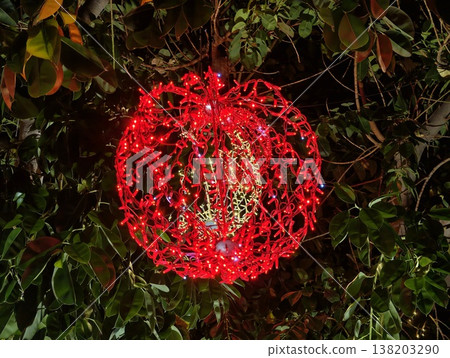 Vivid red globe hangs amidst lush green ivy with detailed wirework and dramatic lighting effects 138203290