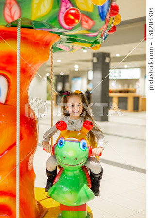 Happy girl riding colorful carousel animal in shopping mall amusement area with bright lights Happy girl riding colorful carousel animal in shopping mall amusement area with bright lights 138203303