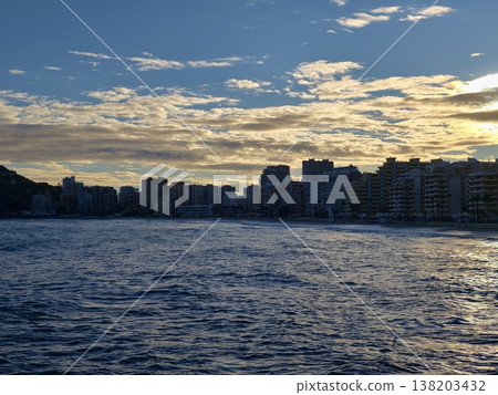Sunset silhouette of urban buildings cast against dramatic clouds over shimmering harbor waters Sunset silhouette of urban buildings cast against dramatic clouds over shimmering harbor waters 138203432