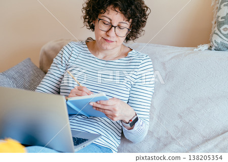 White middle aged woman with curly hair and glasses sits on sofa writing in notebook while using laptop. Home workspace, planning tasks, learning or working remotely in calm bright interior. White middle aged woman with curly hair and glasses sits on sofa writing in notebook while using laptop. Home workspace, planning tasks, learning or working remotely in calm bright interior. 138205354
