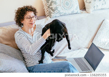 Smiling white middle aged woman with curly hair and glasses sits on sofa holding black cat near laptop. Home interior, relaxed mood, pet interaction, remote work interrupted by daily life moment. Smiling white middle aged woman with curly hair and glasses sits on sofa holding black cat near laptop. Home interior, relaxed mood, pet interaction, remote work interrupted by daily life moment. 138205355