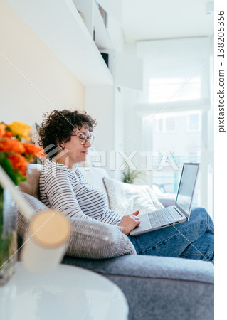 Focused mature woman with curly hair and glasses sits on sofa using laptop. Concentrated expression, natural daylight, calm home environment, remote work, digital lifestyle, quiet focused moment. Focused mature woman with curly hair and glasses sits on sofa using laptop. Concentrated expression, natural daylight, calm home environment, remote work, digital lifestyle, quiet focused moment. 138205356