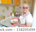 An elderly woman wearing glasses and an apron smiles while holding a bowl of pancake batter and a whisk in her home kitchen. 138205499