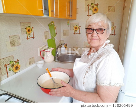 An elderly woman wearing glasses and an apron smiles while holding a bowl of pancake batter and a whisk in her home kitchen. 138205499