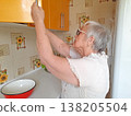 An elderly woman wearing glasses and a white blouse opens a yellow wall cabinet in the kitchen. 138205504
