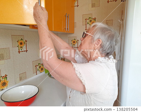 An elderly woman wearing glasses and a white blouse opens a yellow wall cabinet in the kitchen. An elderly woman wearing glasses and a white blouse opens a yellow wall cabinet in the kitchen. 138205504