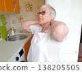 An elderly woman wearing glasses and a white apron stands in the kitchen. 138205505