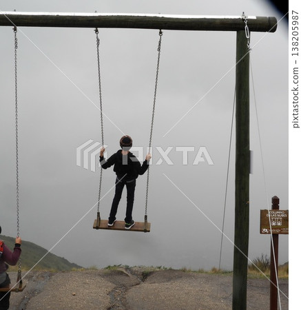 Child standing on a wooden swing suspended over a foggy mountain landscape, viewed from behind. Child standing on a wooden swing suspended over a foggy mountain landscape, viewed from behind. 138205987