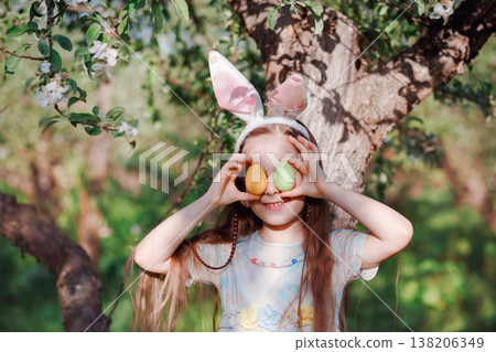 Cute girl wearing bunny ears holding colorful Easter eggs over eyes standing near blooming tree Cute girl wearing bunny ears holding colorful Easter eggs over eyes standing near blooming tree 138206349