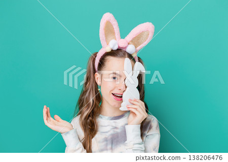 Teenage girl with bunny ears holding wooden bunny toys smiling on bright green background studio 138206476