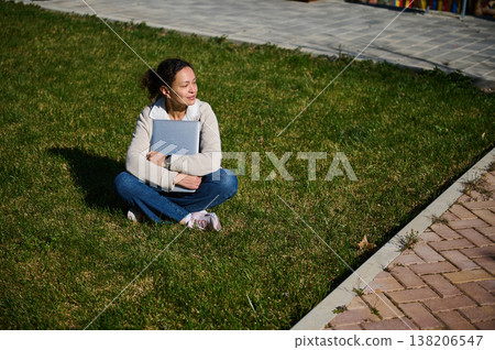 Young Woman Sitting on Grass Hugging Laptop, Relaxed Student Enjoying Sunny Outdoor Moment 138206547