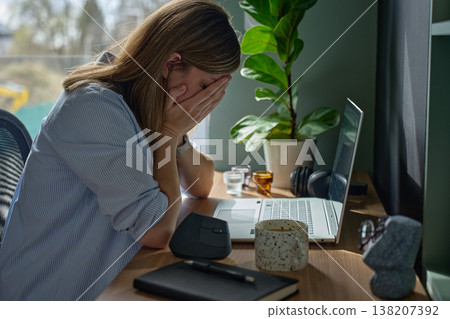 Fatigued woman covering face with hands while sitting at desk with laptop in home office Fatigued woman covering face with hands while sitting at desk with laptop in home office 138207392