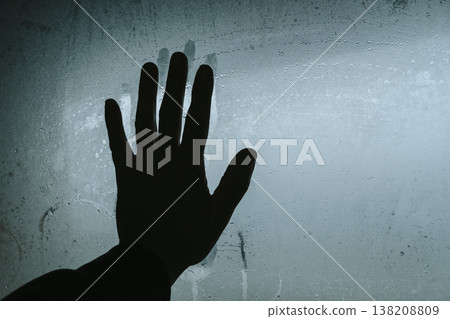 Dark silhouette of a hand pressed against a wet, condensed glass surface in cool blue tones. Conceptual image symbolizing loneliness, separation, or a plea for help through a foggy window with water 138208809