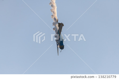 Aerobatic propeller airplane performing a steep dive stunt maneuver while releasing a trail of smoke across a clear blue sky. Aerobatic propeller airplane performing a steep dive stunt maneuver while releasing a trail of smoke across a clear blue sky. 138210779