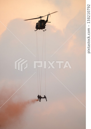 Medics airlift an injured soldier beneath a hovering helicopter using ropes and smoke cover during a rescue demonstration against a sunset sky with clouds at an air show event Medics airlift an injured soldier beneath a hovering helicopter using ropes and smoke cover during a rescue demonstration against a sunset sky with clouds at an air show event 138210792