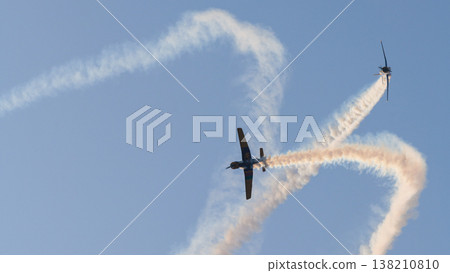 Two aircraft performing aerobatic maneuvers with smoke trails against a clear sky during an aerial aviation display Two aircraft performing aerobatic maneuvers with smoke trails against a clear sky during an aerial aviation display 138210810