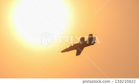 Silhouette of a military training jet aircraft flying across a glowing orange sunset sky during an aerial performance. Dramatic aviation scene against the sun. Silhouette of a military training jet aircraft flying across a glowing orange sunset sky during an aerial performance. Dramatic aviation scene against the sun. 138210822
