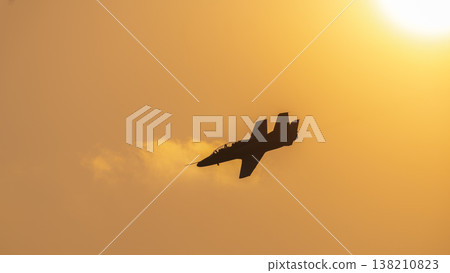 Silhouette of a military training jet aircraft flying across a glowing orange sunset sky during an aerial performance. Dramatic aviation scene against the sun. Silhouette of a military training jet aircraft flying across a glowing orange sunset sky during an aerial performance. Dramatic aviation scene against the sun. 138210823
