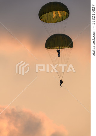 Two combat paratroopers deploying by parachute during an air show demonstration. Silhouettes of troopers descending with glowing parachute canopies against a sunset sky Two combat paratroopers deploying by parachute during an air show demonstration. Silhouettes of troopers descending with glowing parachute canopies against a sunset sky 138210827