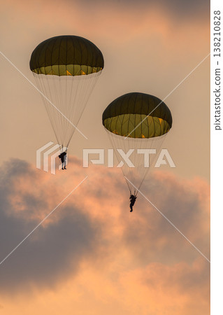Two combat paratroopers deploying by parachute during an air show demonstration. Silhouettes of troopers descending with glowing parachute canopies against a sunset sky 138210828