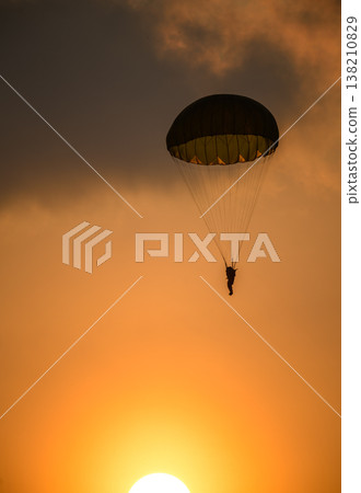 Paratrooper descending in front of a glowing sun. Silhouette of a parachutist against a warm orange sky during an airshow demonstration 138210829