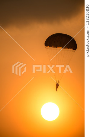 Combat paratrooper deploying a parachute and descending through a glowing orange sunset sky below dark clouds. Silhouette paratrooper during an aerial demonstration 138210830
