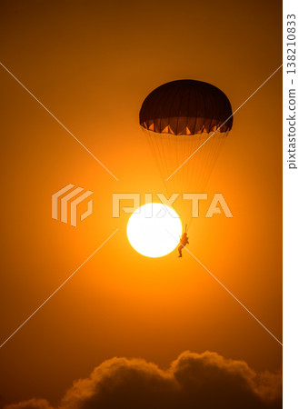 Silhouette of a parachutist descending in front of a glowing sun. A combat paratrooper with a warm orange sky during an airshow demonstration Silhouette of a parachutist descending in front of a glowing sun. A combat paratrooper with a warm orange sky during an airshow demonstration 138210833