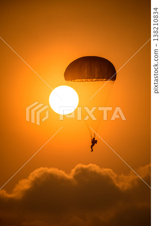 Paratrooper descending in front of a glowing sun. Silhouette of a parachutist against a warm orange sky during an airshow demonstration Paratrooper descending in front of a glowing sun. Silhouette of a parachutist against a warm orange sky during an airshow demonstration 138210834