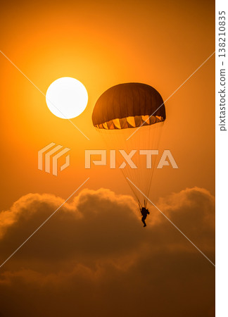 Combat paratrooper deploying a parachute and descending through a glowing orange sunset sky above soft clouds. Silhouette paratrooper during an aerial demonstration Combat paratrooper deploying a parachute and descending through a glowing orange sunset sky above soft clouds. Silhouette paratrooper during an aerial demonstration 138210835