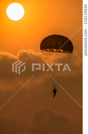 Paratrooper descending in front of a glowing sun. Silhouette of a parachutist against a warm orange sky during an airshow demonstration Paratrooper descending in front of a glowing sun. Silhouette of a parachutist against a warm orange sky during an airshow demonstration 138210836