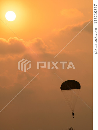 Paratrooper descending in front of a glowing sun. Silhouette of a parachutist against a warm orange sky during an airshow demonstration Paratrooper descending in front of a glowing sun. Silhouette of a parachutist against a warm orange sky during an airshow demonstration 138210837