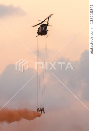 Medics airlift an injured soldier beneath a hovering helicopter using ropes and smoke cover during a rescue demonstration against a sunset sky with clouds at an air show event 138210841