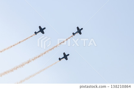 Three aerobatic aircraft performing a synchronized aerial maneuver with curved smoke trails against a clear blue sky during an aviation airshow. Dynamic formation display in an open sky background 138210864