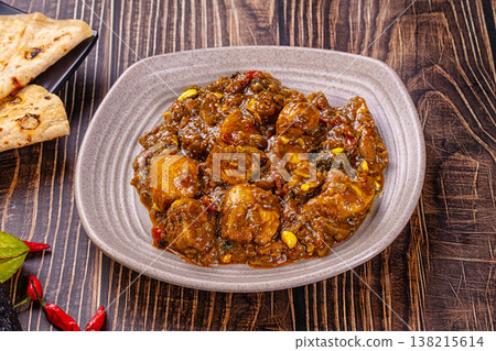 Bowl of traditional spicy chicken curma with flatbread, chilies, and exotic vegetables on a rustic dark wood table. 138215614
