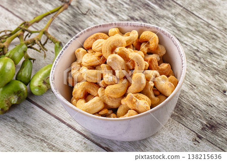 Roasted cashew nuts in a brown bowl with a cluster of raw green cashew fruit on a rustic wooden table. Roasted cashew nuts in a brown bowl with a cluster of raw green cashew fruit on a rustic wooden table. 138215636