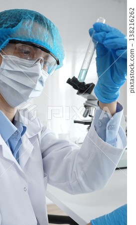 Close up of a scientist in ppe, holding a test tube with blue liquid while conducting research in a laboratory with a microscope. Medicine, health care and science concept Close up of a scientist in ppe, holding a test tube with blue liquid while conducting research in a laboratory with a microscope. Medicine, health care and science concept 138216262